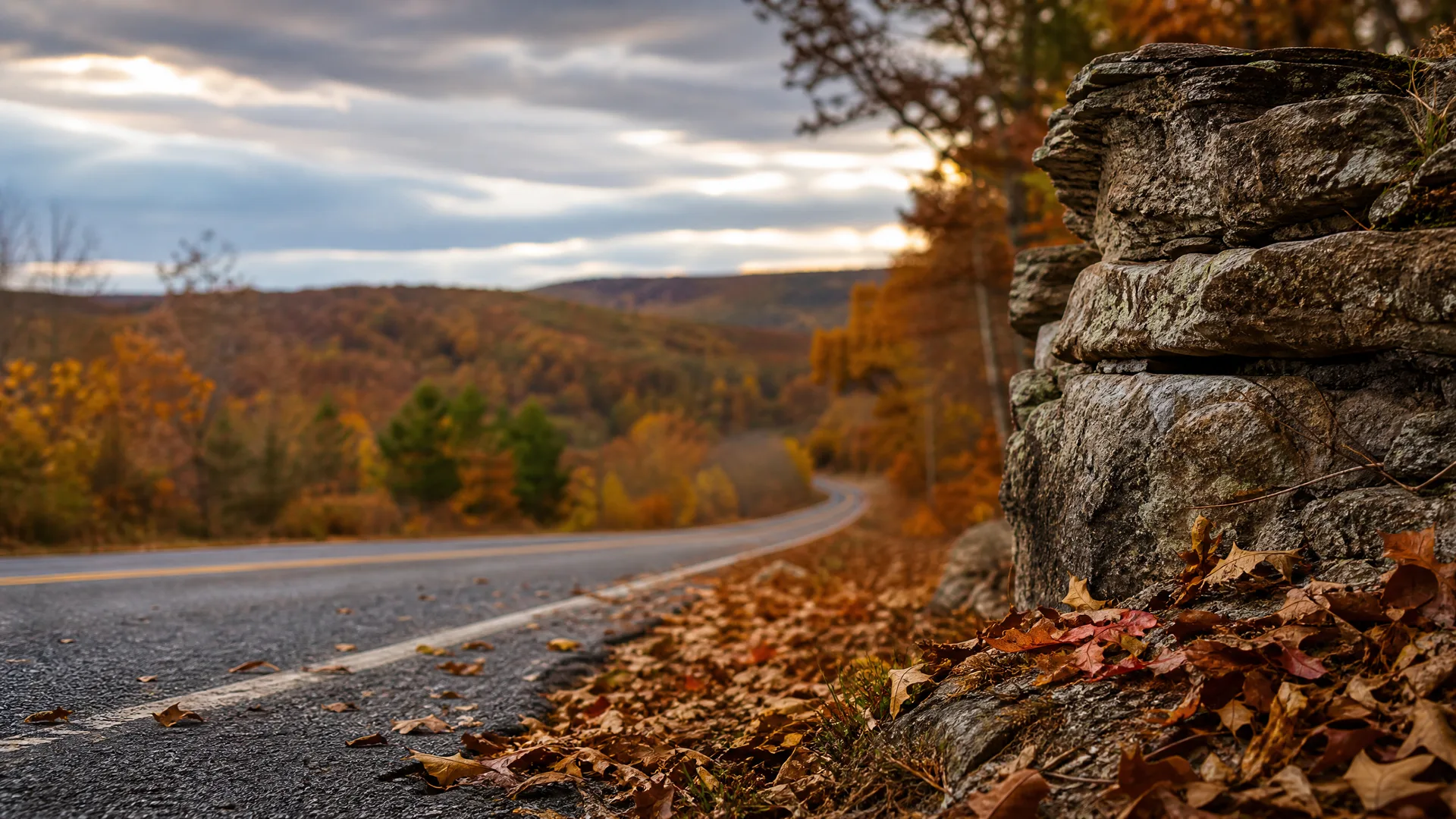 A curving two-lane road with painted edge lines runs past a stacked rock outcrop and fallen leaves, with autumn-colored forested hills and a cloudy sky in the blurred background.